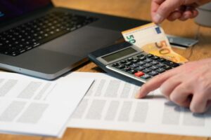 Close-up of hands using calculator with euro notes and laptop on desk.